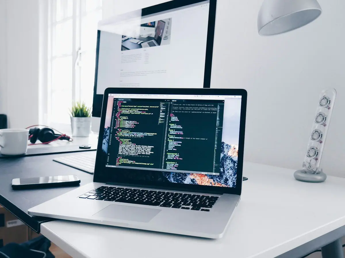 Heller Arbeitsplatz mit aufgeklapptem Laptop, auf dem Bildschirm Programmcode, daneben Tasse und Pflanze